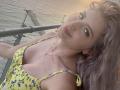 EliseMonet with pink hair, yellow floral dress, and gold necklace, showing tits, pouty face, and a slight smile, on a boat. Water and metal railing in the background.