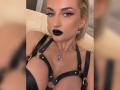 MademoiselleBella with black leather harness, large tits, black lipstick, dramatic eyeliner, and diamond earrings, looking seductively at the camera. Blond hair, fair skin, and a silver cross necklace.