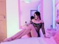 MelisaDaltonn est assise sur un lit, les jambes croisées, vêtue d'une robe noire en résille, qui expose ses seins et ses parties intimes, avec de longs cheveux noirs, et un éclairage néon rose en arrière-plan..