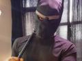 LilithAndAsmodeo in a black mask with eyes visible, holding a black stick, wearing a dark purple shirt, with a white band across the forehead.