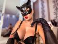 Mademoiselle Bella, con maschera da gatto nera, rossetto rosso, reggiseno di pelle nera, grandi seni, guanti neri, braccio tatuato, in una camera da letto, che guarda la fotocamera con un'espressione sicura..