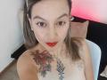 ArianaBlonde with red lipstick, tattoos on her chest and shoulder, brown hair in puffs, looking at camera, topless, small earrings, white chair, black and pink pillows.
