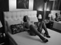AmmyFrancaise is lying on a bed, topless with big tits, wearing black lace stockings and high heels, eyes closed, arms behind head, legs spread, in a black-and-white photo.