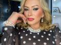MisseScarleth with blonde curls, wearing a black polka dot top, pearl necklace, and earrings, has pink lipstick and blue eyeshadow, touching her cheek with a pearl bracelet.