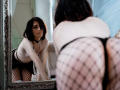 ReginaMay with short black hair, white top, black thong, and fishnet stockings, bending over, showing her ass in a mirror, looking at herself with a serious expression.
