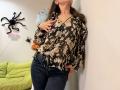 Tilliana, com uma blusa floral marrom, mangas bufantes, jeans escuros e unhas rosa, apoiada em uma parede branca, com a mão no peito, a cabeça inclinada para cima, cabelo castanho, sem seios ou nádegas visíveis..