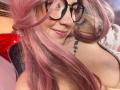 EmmaFeri with pink hair, glasses, and a smile, showing a tattoo on her right shoulder, with her bare boobs slightly visible. She's wearing a pink hairpin.