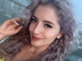 EliseMonet with curly, purple-tinted hair, smiling with red lipstick, hand in hair, wearing a yellow top, white nails, and a gold necklace.