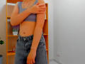 Steisii has tan skin, small tits in a blue bra, and a denim skirt, covering one arm with a hand, wearing a bracelet, standing in a room.