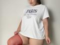 Madrinia with medium tits, wearing a white t-shirt and red thigh-high stockings, slightly lifting shirt to reveal bare ass, standing against a plain wall.