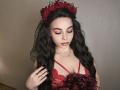 Niferttiti with long black wavy hair, wearing a red lace dress, flower crown, and veil, looking down with closed eyes, showing her small tits, and black nail polish.