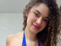 SanetPerlas with curly brown hair, light skin, red lipstick, small zits on cheeks, wearing a blue halter top, smiling slightly, showing bare shoulders.