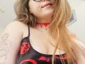 KarKutie with long brown hair, glasses, red choker, black tank top with red skulls, tattoos on arms, showing her tits, smiling slightly, in a casual selfie.