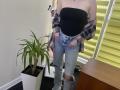 Nesie with pale skin, black strapless top, pink plaid shirt off-shoulder, ripped blue jeans, and grey boots, standing next to a potted plant and yellow blinds.