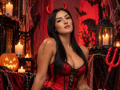 AriiaMon with long black hair, red devil horns, red corset showing big tits, holding a red pitchfork, surrounded by candles and a pumpkin, in a sexy Halloween setup.
