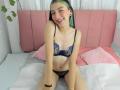 AbbyHillsI with green-tipped hair, smiling, wearing a blue lace bra and matching panties, sitting on a bed with pink pillows, showing her small tits and cute pussy.