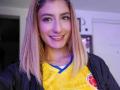 Kaiix has blonde hair, a nose ring, and a smile. She's wearing a yellow soccer jersey with a red logo and blue trim, standing in a room.
