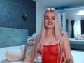 CoquineElise, avec de longs cheveux blonds, un haut rouge transparent qui révèle ses petites seins, souriante, portant une croix en pendentif, un tatouage sur le bras droit, assise sur un lit avec un animal en peluche blanc..