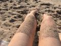 Avellia's sandy-covered thighs and calves, with her bare feet in the sand, showing a beach scene from a first-person perspective. Her skin is light and slightly sunburned.