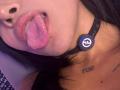 MajoJones's face, tongue out, with black lace choker and Razer logo, small black nose, pink tongue, and tattooed neck. Boob tattoo visible. MajoJones has dark hair.