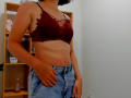Steisii has small tits, wearing a red lace bra, and blue denim shorts, standing with her right hand on her hip, in a room with a wooden shelf.