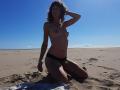 Topless Aiyana with small tits, black panties, kneeling on sandy beach, hand in curly brown hair, clear blue sky, sunlight, visible tan lines, beach towel beneath her.