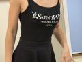 Rorria's black tank top and skirt show her small tits, with a tight fit highlighting her slim, tan-skinned body and subtle arm muscles. She's in a simple indoor setting.