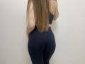 Palerianna stands with her back to the camera, showcasing long, straight brown hair that reaches down to her lower back. She wears a black sports bra and tight, dark blue leggings that highlight her curvy, well-toned buttocks. The lighting is even, with no shadows, and the image has a simple, clean composition. The focus is on Palerianna's lower back and buttocks, emphasizing her fit, hourglass shape. The texture of her leggings is smooth, contrasting with the softness of her hair.