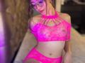 AprilBoo en lingerie en dentelle rose, montrant ses seins à travers un haut en filet, avec un short assorti, portant des lunettes et un collier, assise sur un lit..