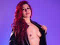 AntonelaRiveiro with red curly hair, black leather jacket open, exposing her tits with nipple piercings, tattoo under left breast, against purple background, looking at camera with slight smile.