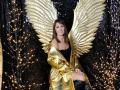 MiraMixi with golden wings, off-shoulder black top, shiny gold coat, and black thigh-highs, sitting with a subtle smile, surrounded by glowing string lights.