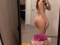 EarthAmspaugh in beige thong and crop top, showing her ass in a mirror selfie, holding a phone, with a pink bag of purple flowers in the foreground.