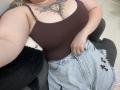 DiLaria's brown tank top shows her large tits with a sun tattoo above her cleavage. She's wearing light blue, frayed denim shorts, sitting on a dark gray couch.