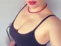 AvaQueen with red bead necklace, black ribbed tank top, showing her big tits and pink lipstick, slight armpit visible, standing against a light grey wall.