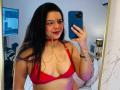 ViolettaRouse with red bra, chains, and choker, holding phone, smiling, long black hair, red lipstick, medium tits, mirror selfie, tattoo on left wrist, bed in background.