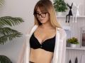 ChloeKingsley with blonde hair, black lace bra, and white shirt, wearing black glasses, looking down, showing her small tits, in a room with green plants and white shelves.