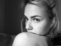 ArnettaNazario's face in a black-and-white photo, eyes looking back, arms crossed over bare shoulders, short blonde hair with a hairpin, subtle makeup, slightly parted lips.