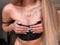 Karish with long blonde hair, covering her tits with hands, black corset, white nails, tattoos on chest, necklace, rings, and pink panties, standing in a room.