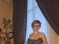 SabrinaShine with short, dirty-blonde hair, black glasses, and a sheer gray lace top showing her small tits. She's smiling, standing in front of a window with curtains.