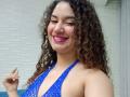 SanetPerlas with curly brown hair, pink lipstick, and blue halter top with cut-out, smiling at camera, showing small tits, pointing with right index finger.