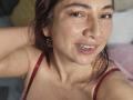 AlessiaRed has freckled skin, brown eyes, and a slight smile, wearing a red strap, showing her tits, with brown hair and colorful earrings, in a selfie.