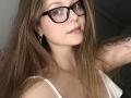 KarKutie with long brown hair, black glasses, pink lips, small nose, white tank top, showing her tits, arm tattoo, and silver earrings, looking at camera, smiling slightly.