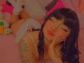 AnnieGrey lying on a pink bed, eyes closed, wearing white lingerie, showing her ass, black braids, white socks, choker, and a floral tattoo on her left arm.