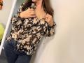 Tilliana, com cabelo castanho, segurando aberta uma blusa preta com estampas florais, expondo seus seios, vestindo jeans azuis, em pé em frente a uma parede branca..