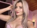 EliseMonet, avec ses cheveux blonds, son rouge à lèvres rose et son sourire charmant, montre ses seins dans un soutien-gorge en dentelle rose, une main dans ses cheveux, un plafond en bois et un maquillage discret..