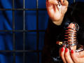NenaBlack's hand with red nail polish holds a key while wearing a shiny metal butt plug. Dark blue background, black grid pattern. NenaBlack's finger is slightly curled.