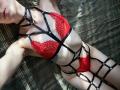 GwynBartell allongée sur un lit, portant un soutien-gorge et une culotte en dentelle rouge, avec un harnais noir à bretelles, exposant ses seins et ses parties intimes, peau pâle, tatouage sur le côté droit, collier ras du cou..