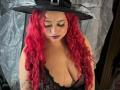 Estefy69 with red curly hair, black lace lingerie, and a witch hat, looking down with dark makeup, showing her large boobs, tattoos, and a nose ring.