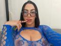 CamilleJuicyHot with blue lace top, big boobs, finger in mouth, glasses, black hair, tattoo under boobs, white background, looking at camera.