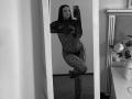 Olieri with long dark hair, wearing a sheer black lace bodysuit, standing on one leg, taking a mirror selfie, showing her tits and pussy through the lace. Black and white.